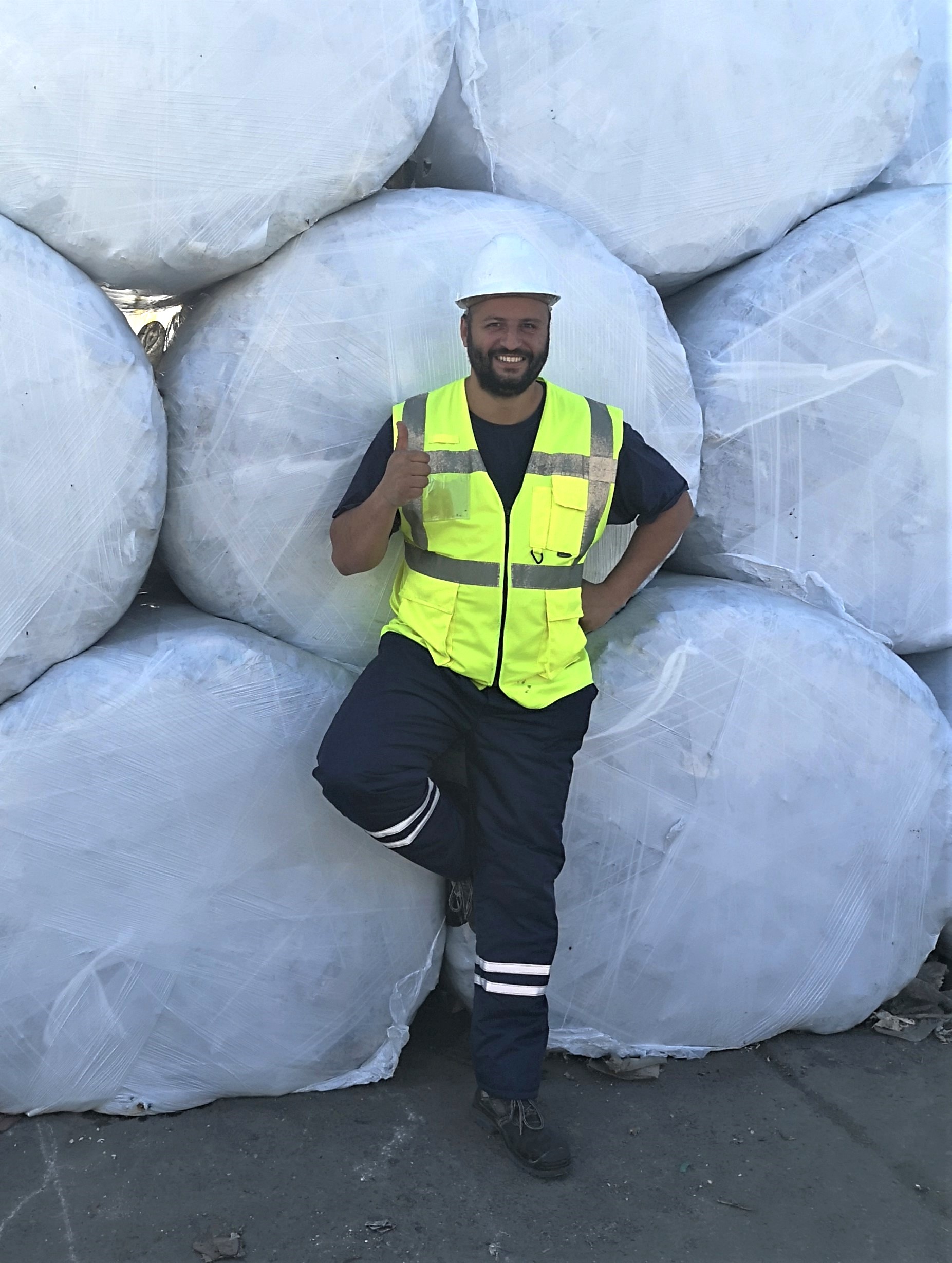 A person in a yellow safety vest posing with a "thumb's up" in front of stacked round bales, working on waste management