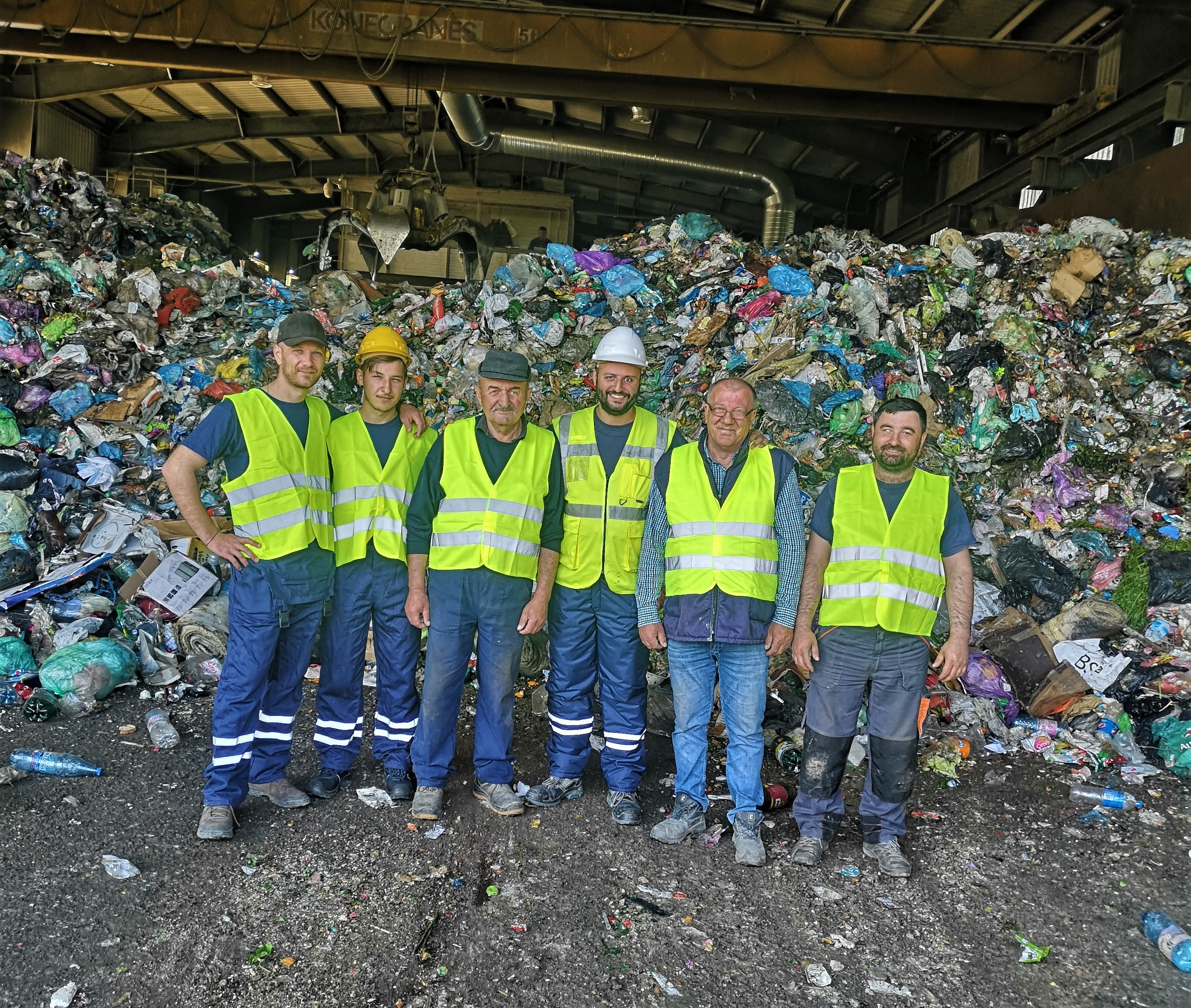 Six people in yellow safety vests posing in front of a tall pile of household, working on waste management