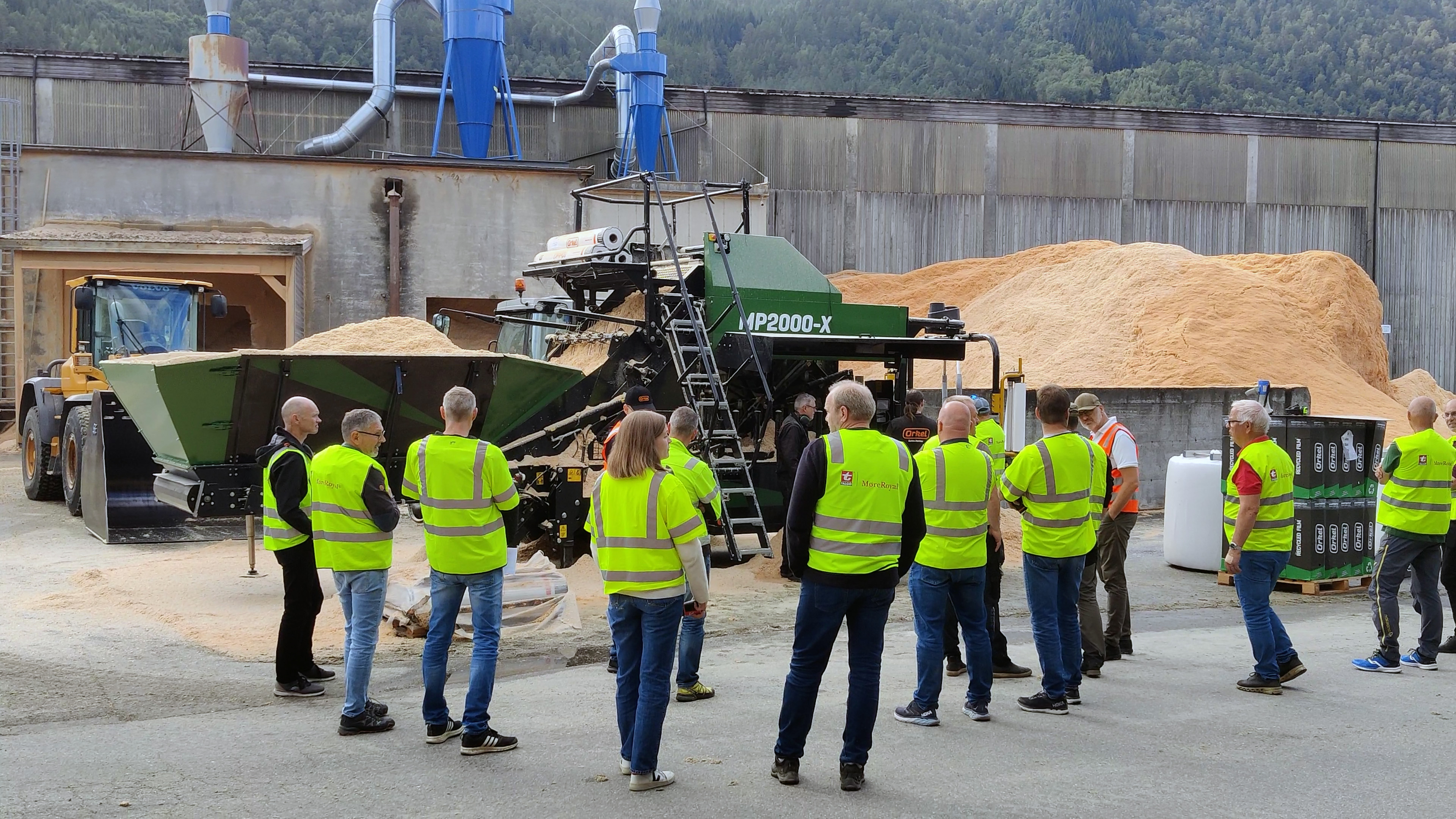 A crowd of people wearing yellow safety vests standing in front of an MP2000-X baling wood shavings, with a large pile of wood shavings in the background.