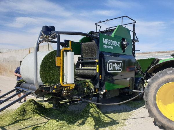 A Orkel MP2000-X compactor with a round bale of grass lying on the wrapping table, being wrapped in recycled film