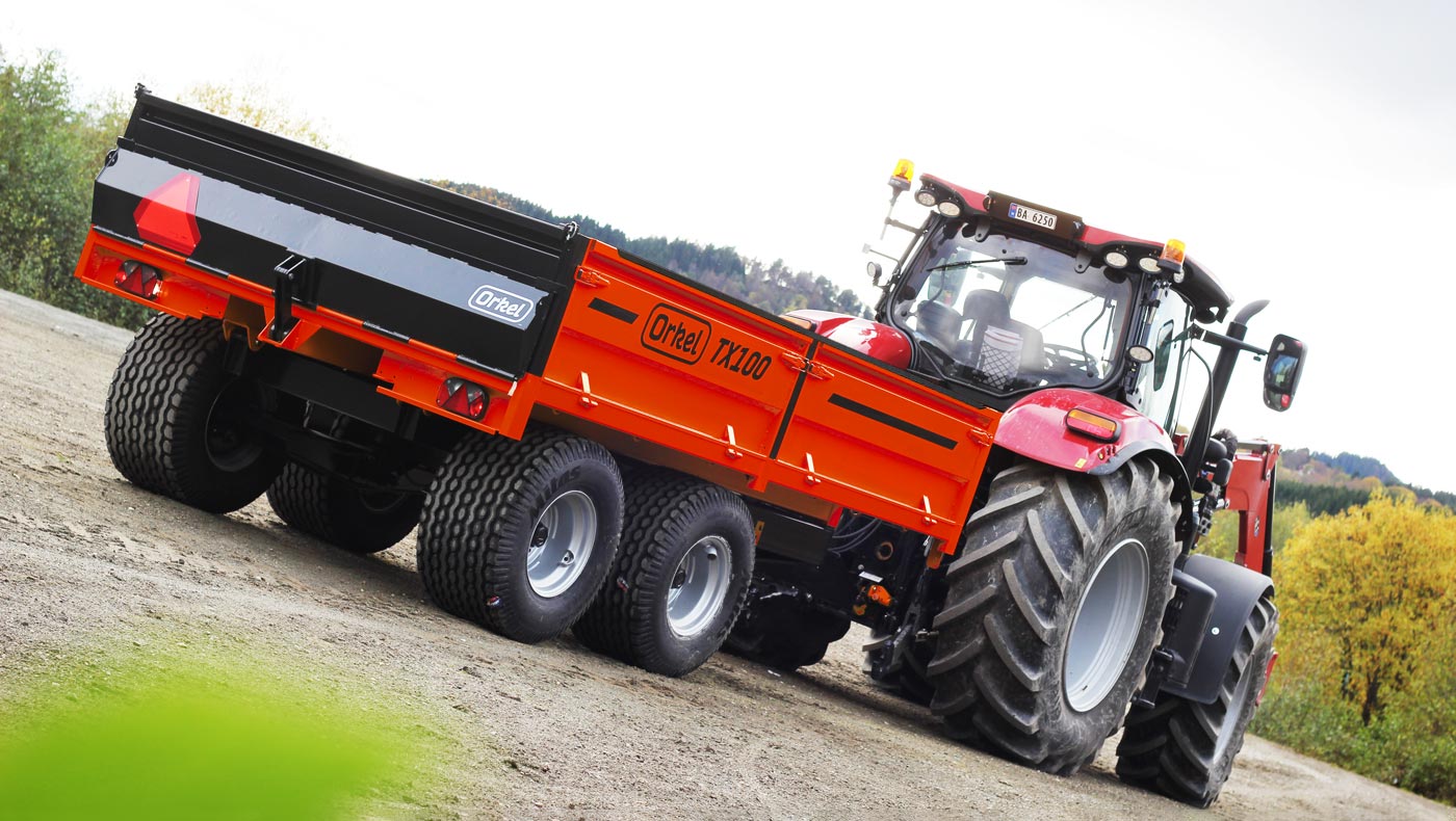 Rød Orkel TX100 traktorhenger stående ute. Trær og blå himmel i bakgrunn