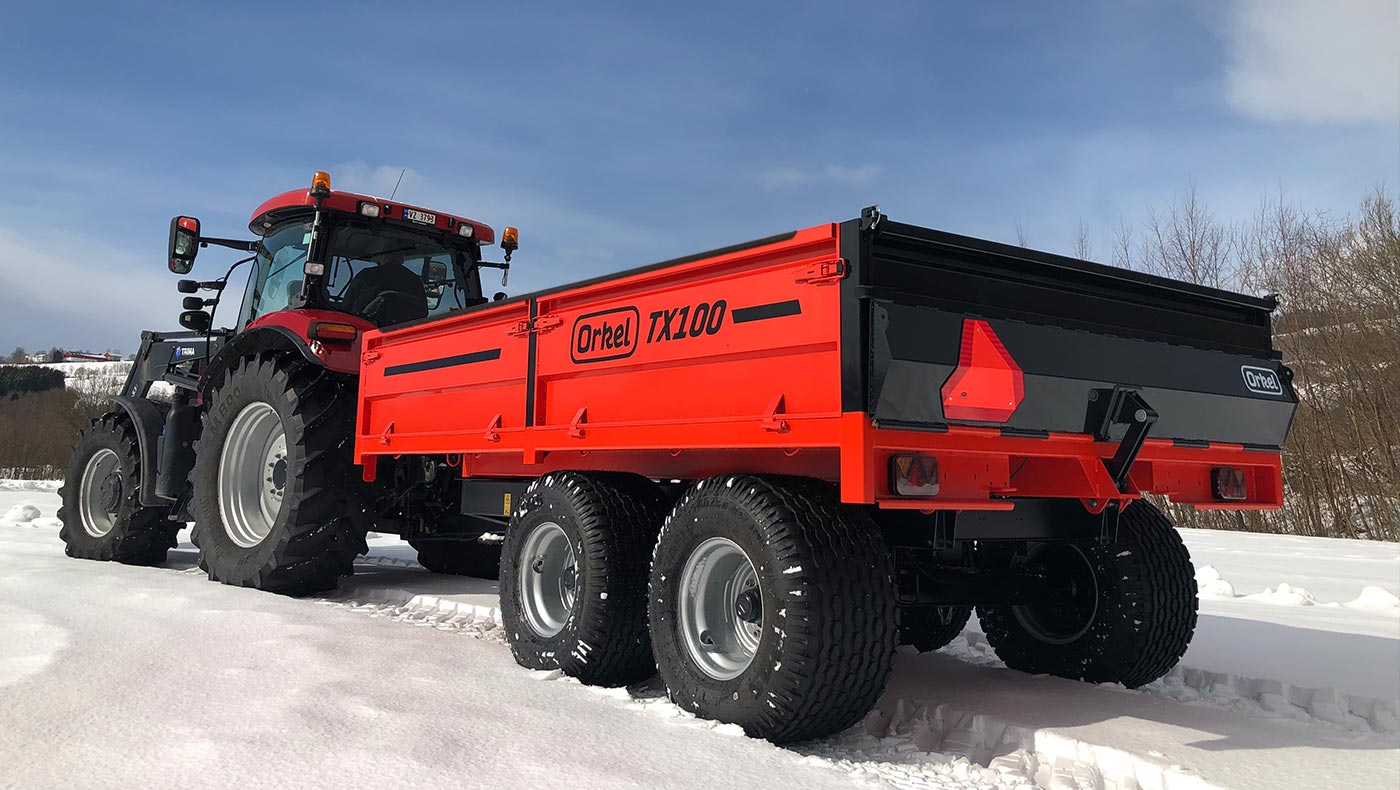 Rød Orkel TX100 traktorhenger stående ute i snølandskap. Trær og blå himmel i bakgrunn
